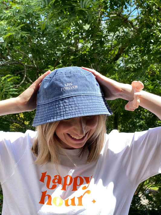 Person wearing a white t-shirt with 'happy hour' text and a blue denim bucket hat embroidered with the slogan 'live forever'