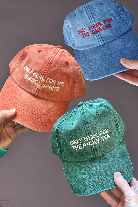 Three colourful baseball caps with humorous custom text held by hands against a grey background.