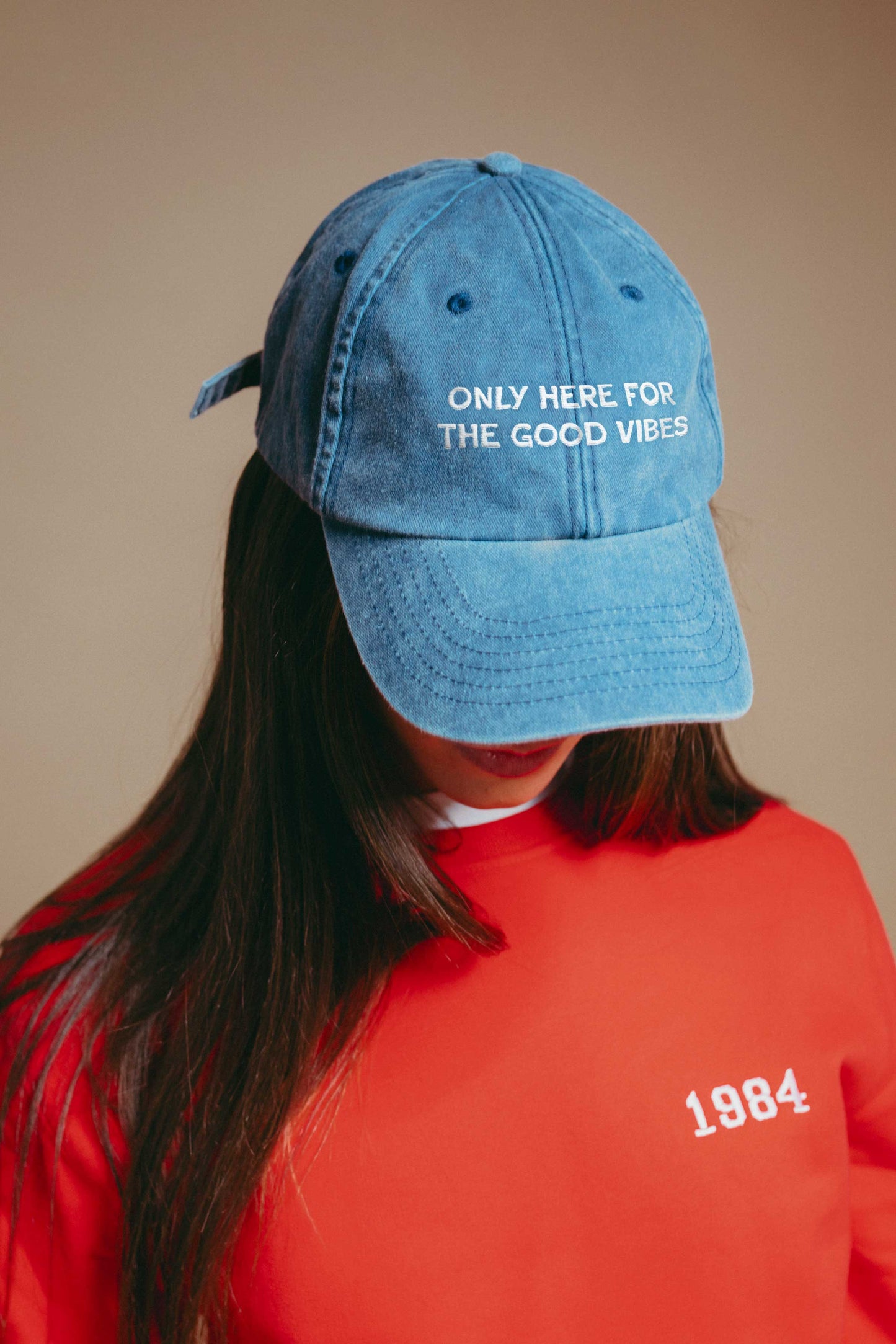 Person wearing a blue denim cap with text 'only here for the good vibes' and a red sweatshirt with '1984' on a beige background