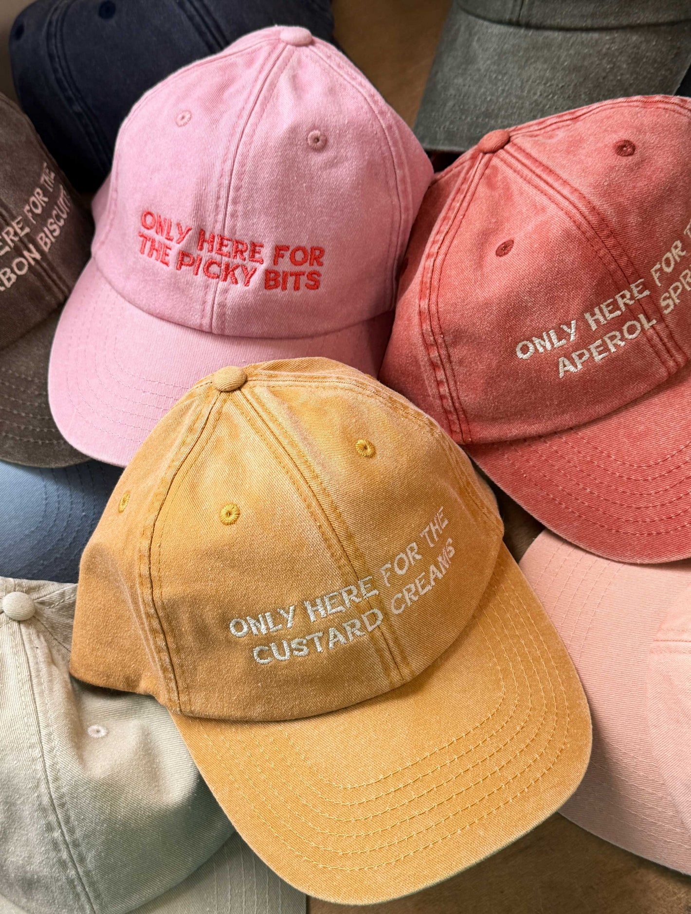 Four colourful baseball caps with funny 'only here for...' slogan text designs on a wooden surface.