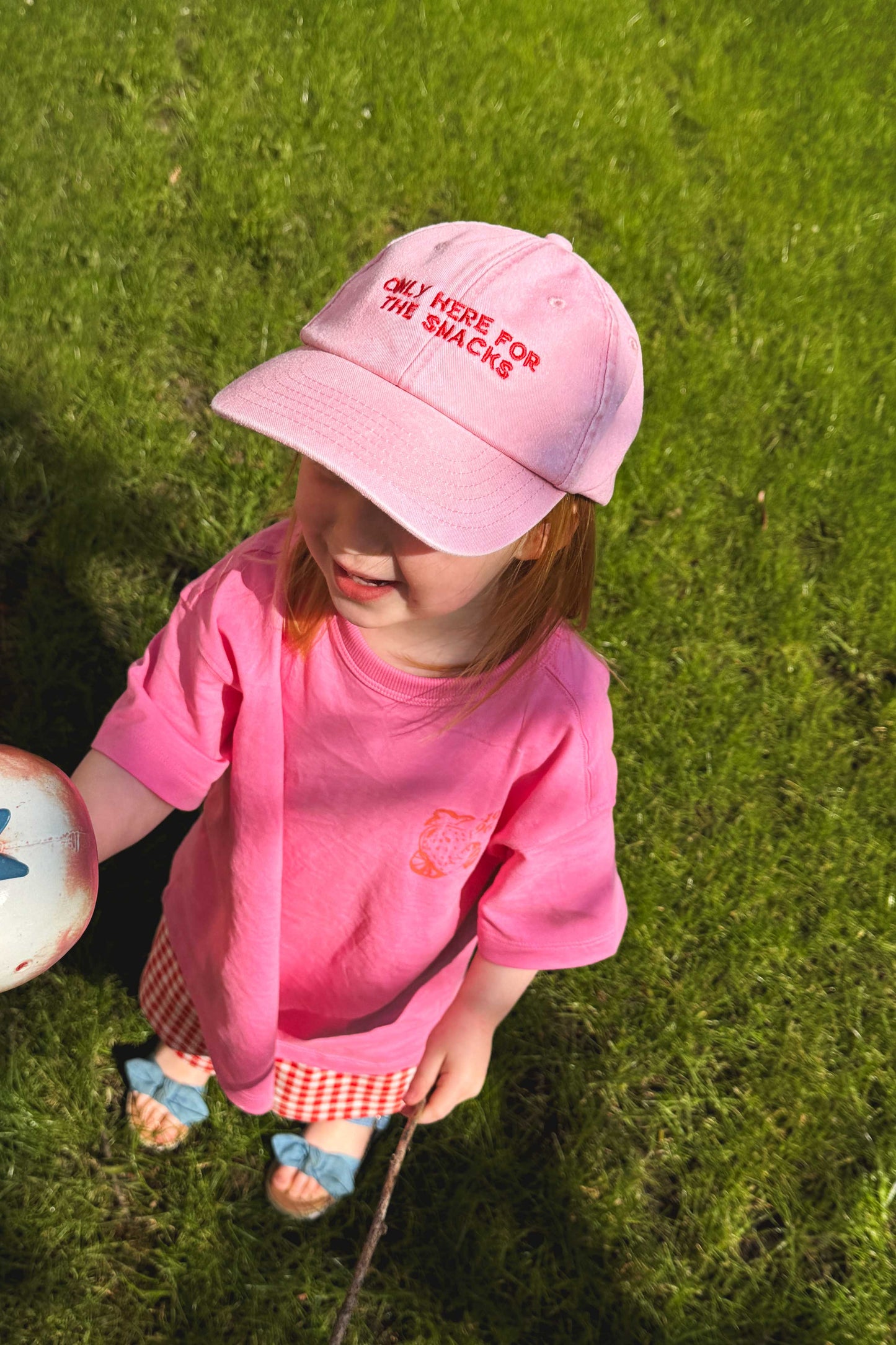 Child wearing a pink cap with the slogan 'only here for the snacks' while stood on gras