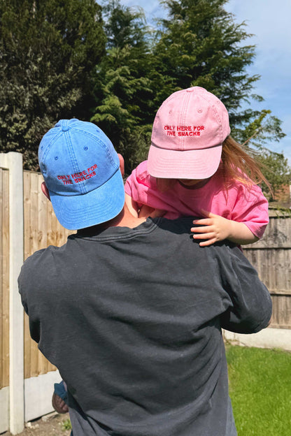 Person holding a child wearing pink cap with text, both outdoors with trees in the background