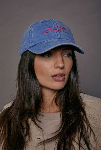 Woman wearing a blue denim cap with pink text 'only here for the tea' against a grey background