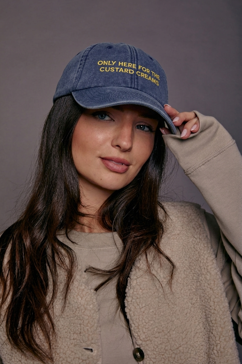 Woman wearing a denim cap with yellow text 'only here for the custard creams', posing against a grey background