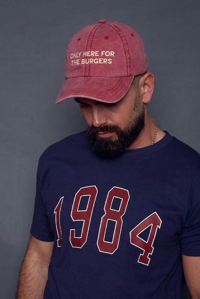 Man wearing a navy blue t-shirt with '1984' and a red cap with text 'only here for the burgers' on a grey background