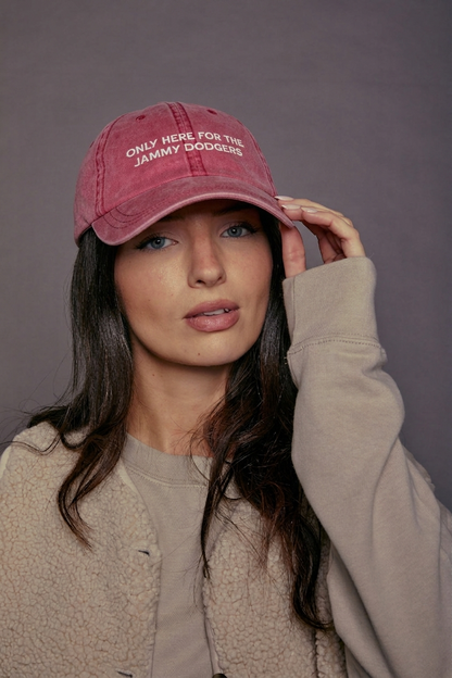 Woman wearing a faded red cap with text 'only here for the jammy dodgers' posing against a grey background