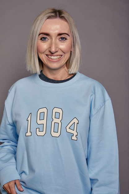 Person wearing a light blue, oversized sweatshirt with '1984' printed on it against a plain background