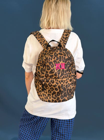 A person viewed from behind wearing a leopard-print backpack with a bright pink, personalised monogram initial on the back, over a white t-shirt and blue checkered pants, against a solid dark blue background.