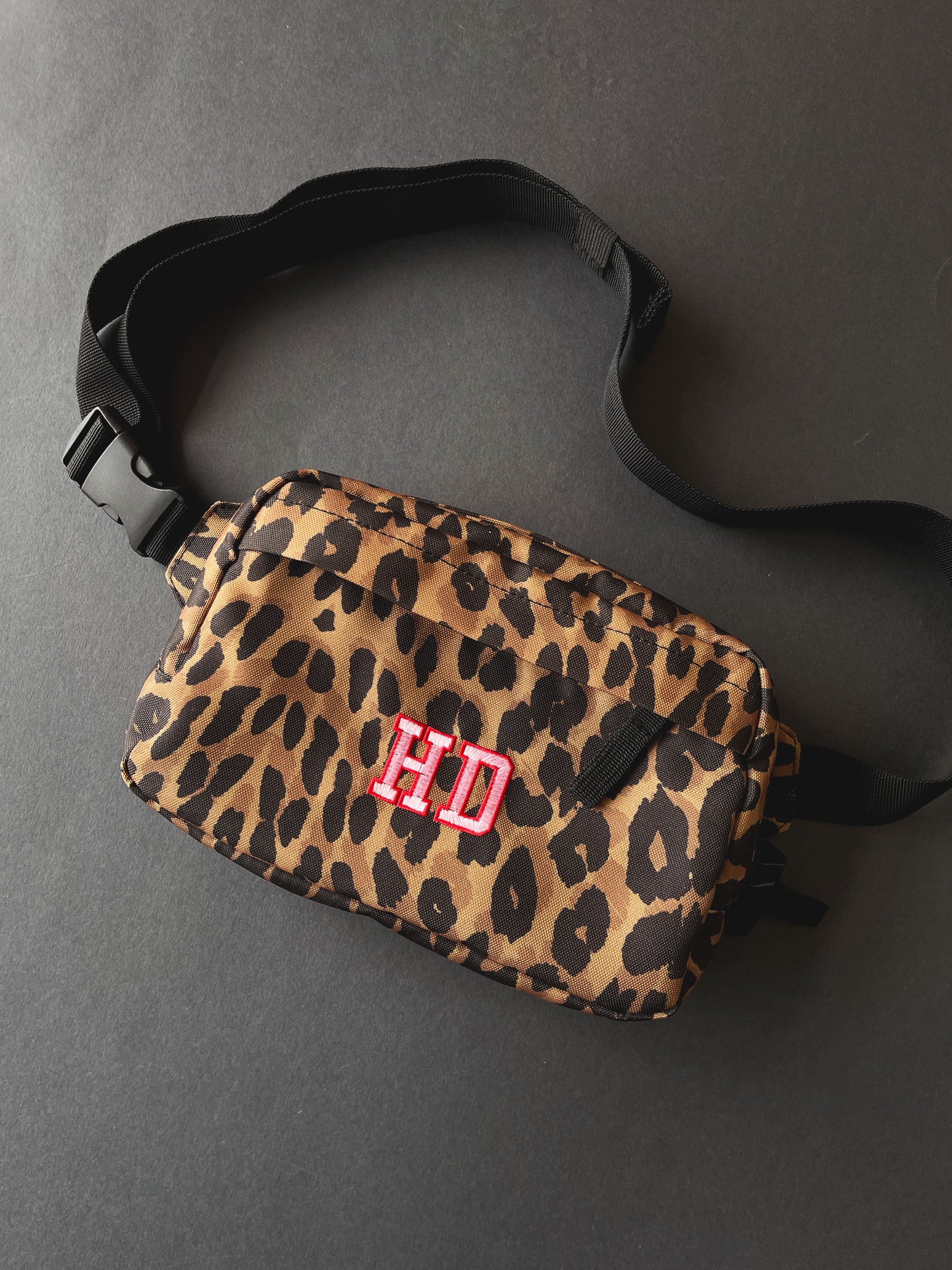 A leopard print crossbody bag with a black adjustable strap and a pink and red embroidered 'HD' monogram on the front, shown on a dark grey background.
