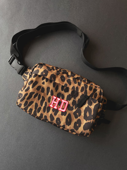 A leopard print crossbody bag with a black adjustable strap and a pink and red embroidered 'HD' monogram on the front, shown on a dark grey background.