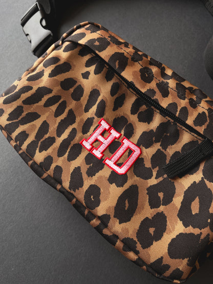 A close-up of a leopard print pouch bag with a black zipper, black strap, and a red embroidered 'HD' monogram on the front, photographed on a dark surface.