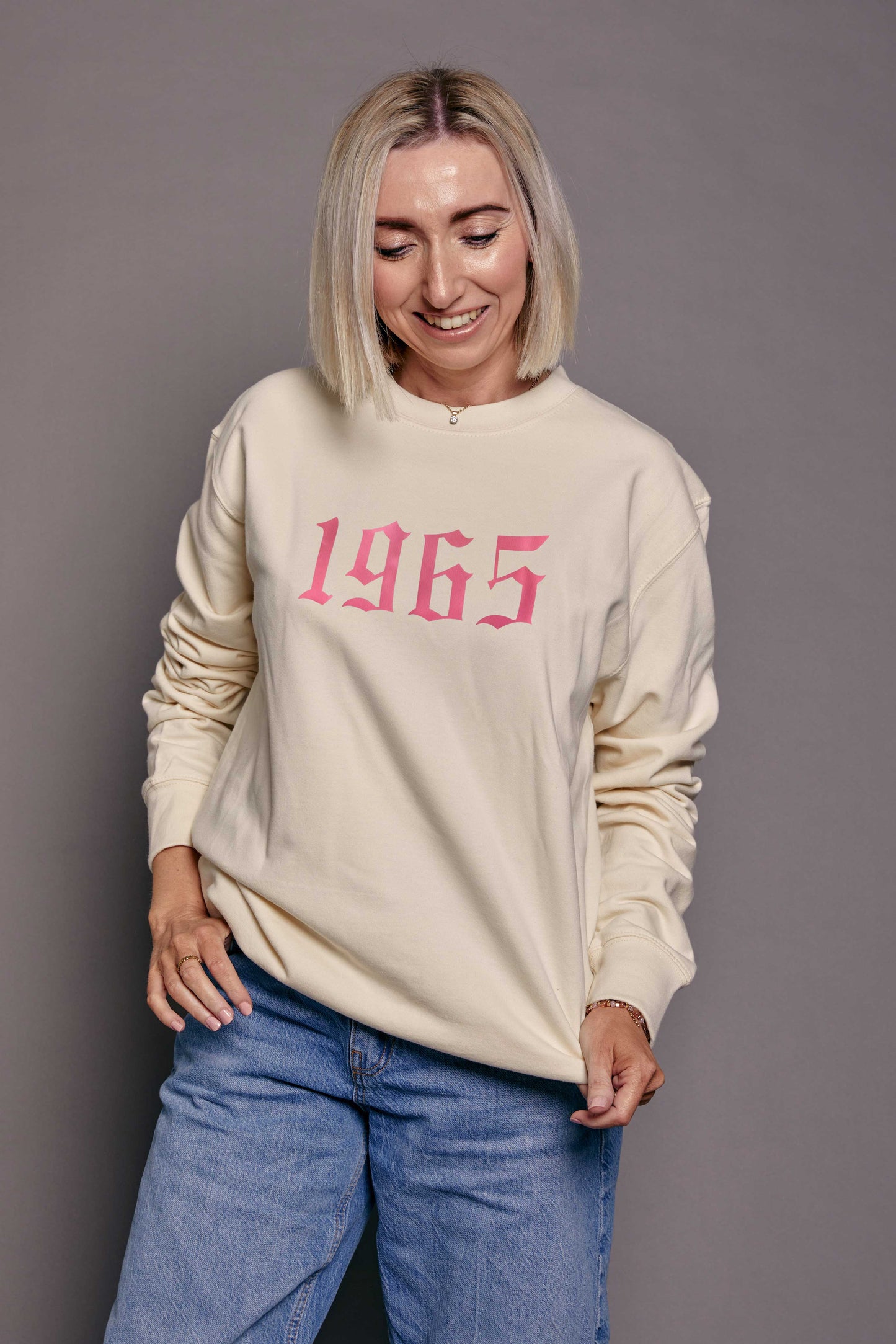 Person wearing a cream sweatshirt with '1965' printed in pink on a grey background