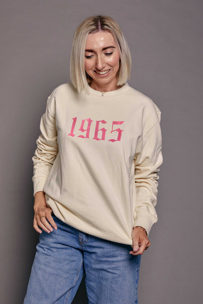 Person wearing a cream sweatshirt with '1965' printed in pink on a grey background