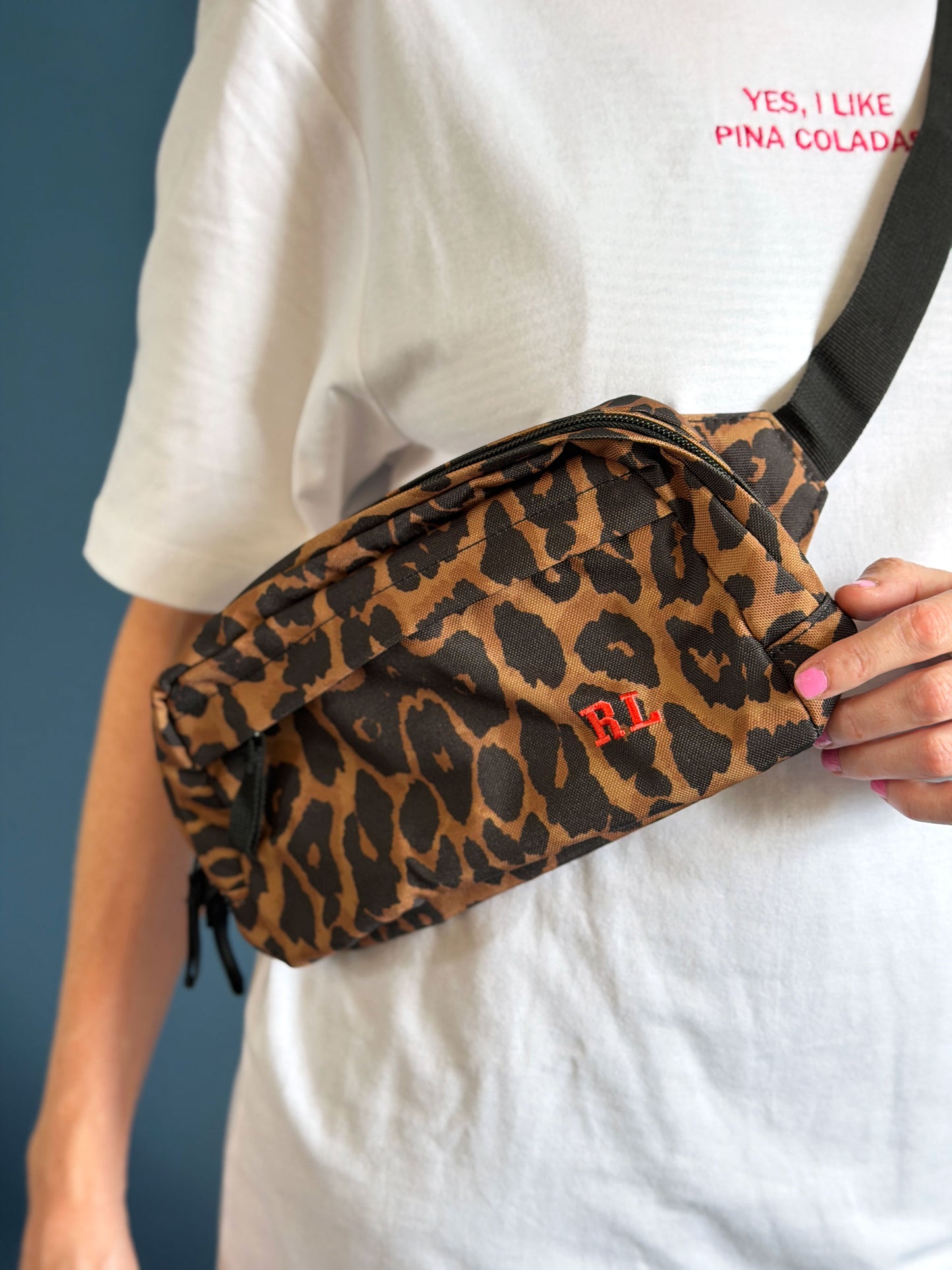 A person wearing a white t-shirt with pink embroidered text and holding a leopard print crossbody bag with red embroidered 'RL' initials. The background is a solid dark blue wall.