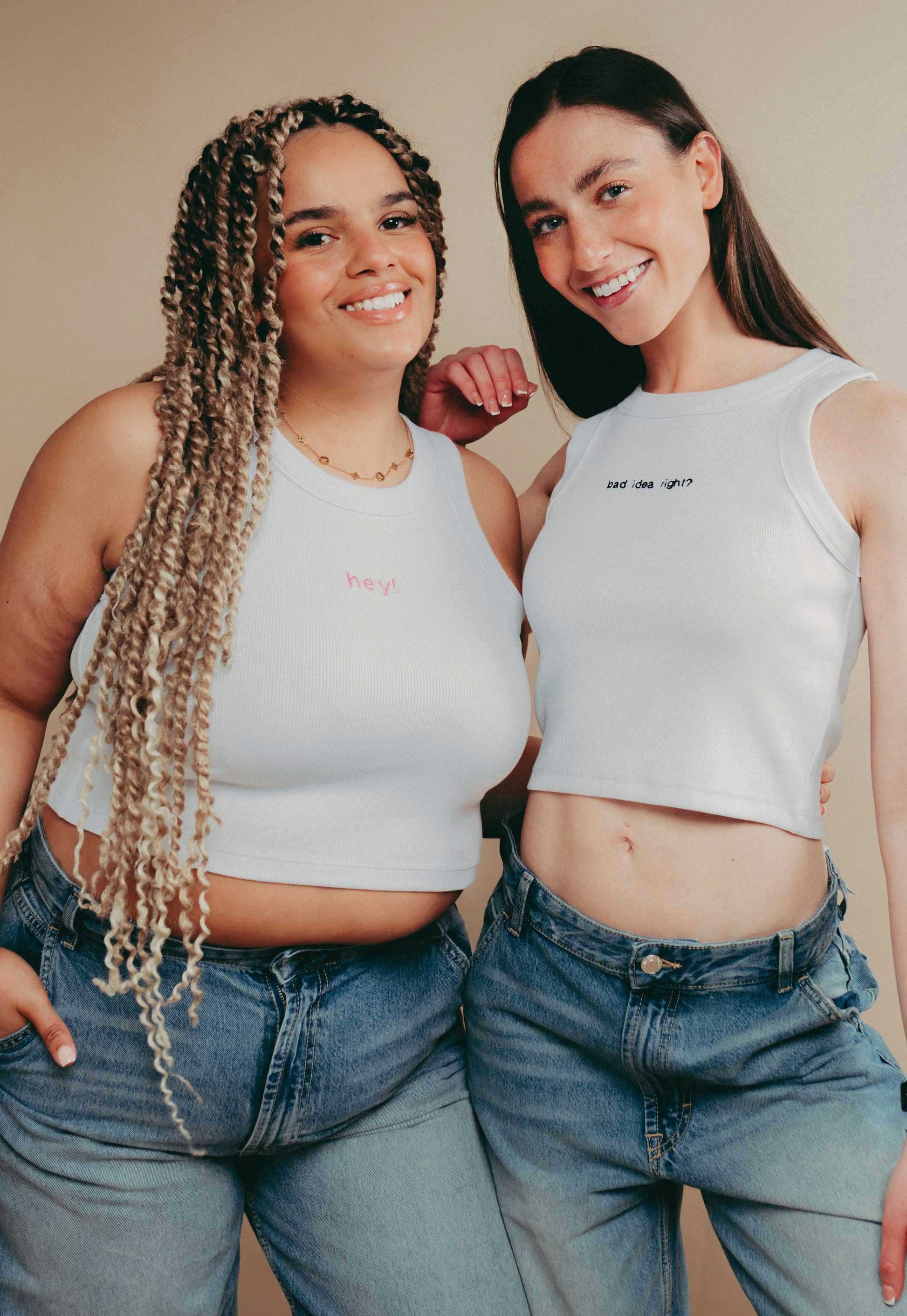 Two women wearing white slogan crop top vests and blue jeans standing against a beige background