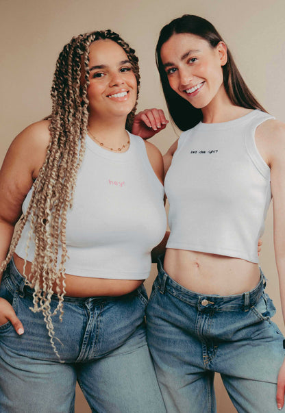 Two women wearing white slogan crop top vests and blue jeans standing against a beige background