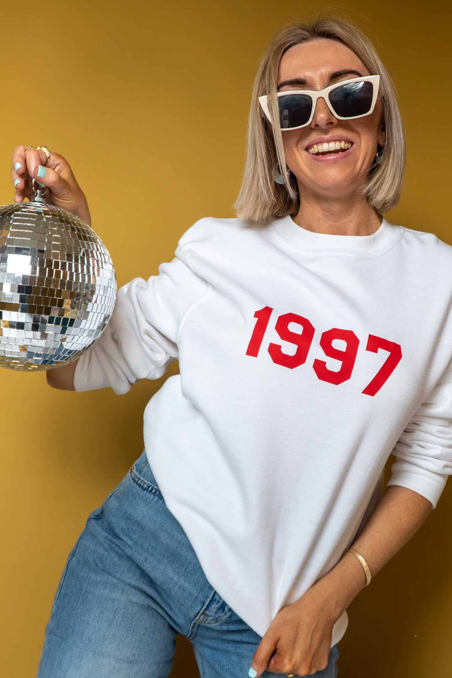 Person wearing a white sweatshirt with '1997' on it, holding a disco ball against a yellow background.
