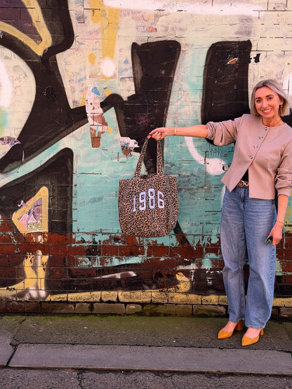 Woman holding a tote bag with '1986' printed on it, standing in front of a graffiti-covered wall.
