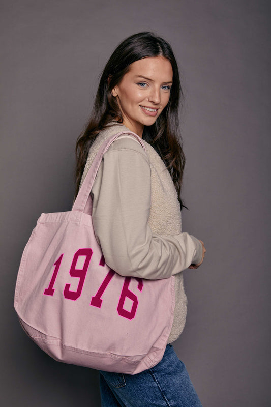 Woman holding a pink tote bag with '1976' printed on it against a grey background