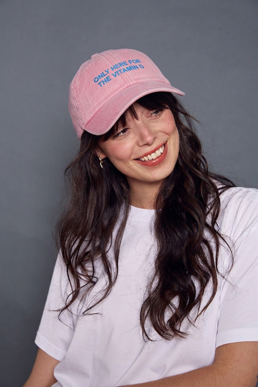 Woman wearing a pink cap with blue text 'only here for the vitamin d' against a grey background