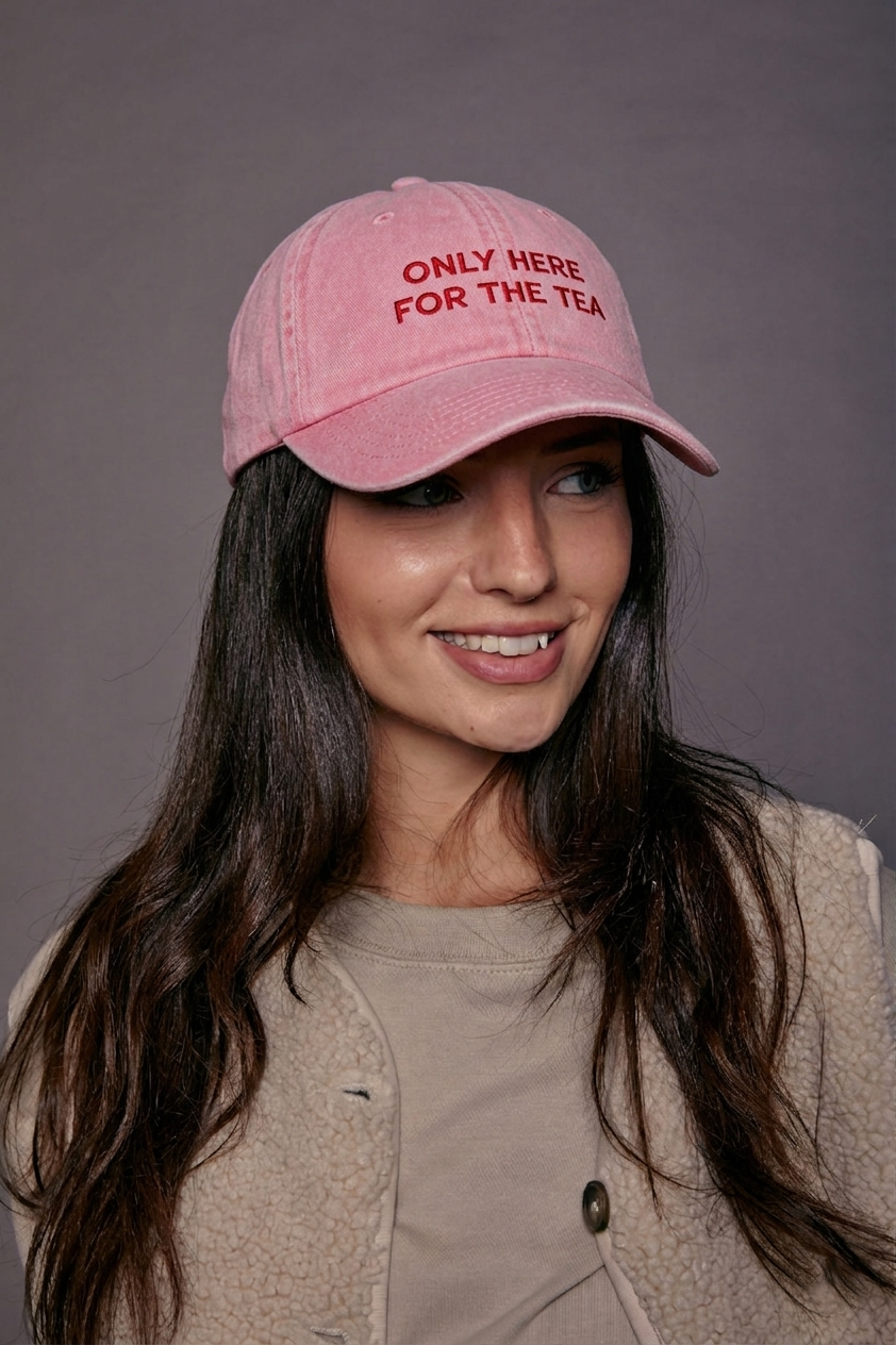 Woman wearing a pink cap with text 'only here for the tea', smiling against a gray background