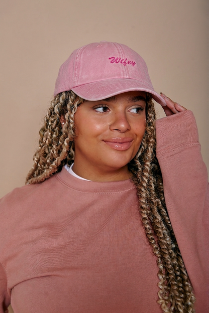 Person wearing a pink cap with 'Wifey' embroidery and matching pink sweatshirt against a beige background
