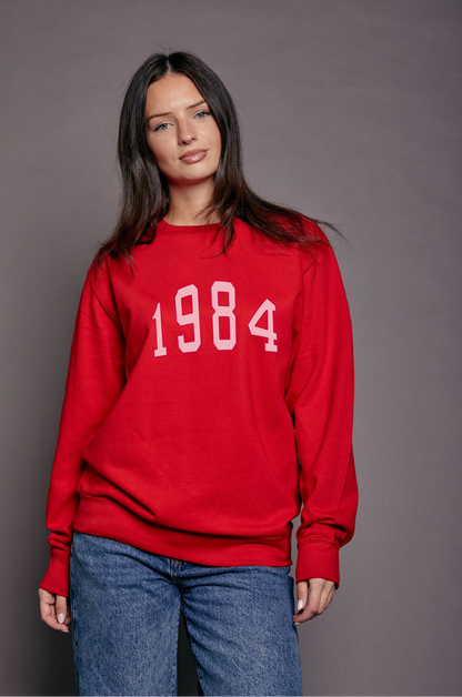 Person wearing a red sweatshirt with '1984' printed on it in pink against a grey background