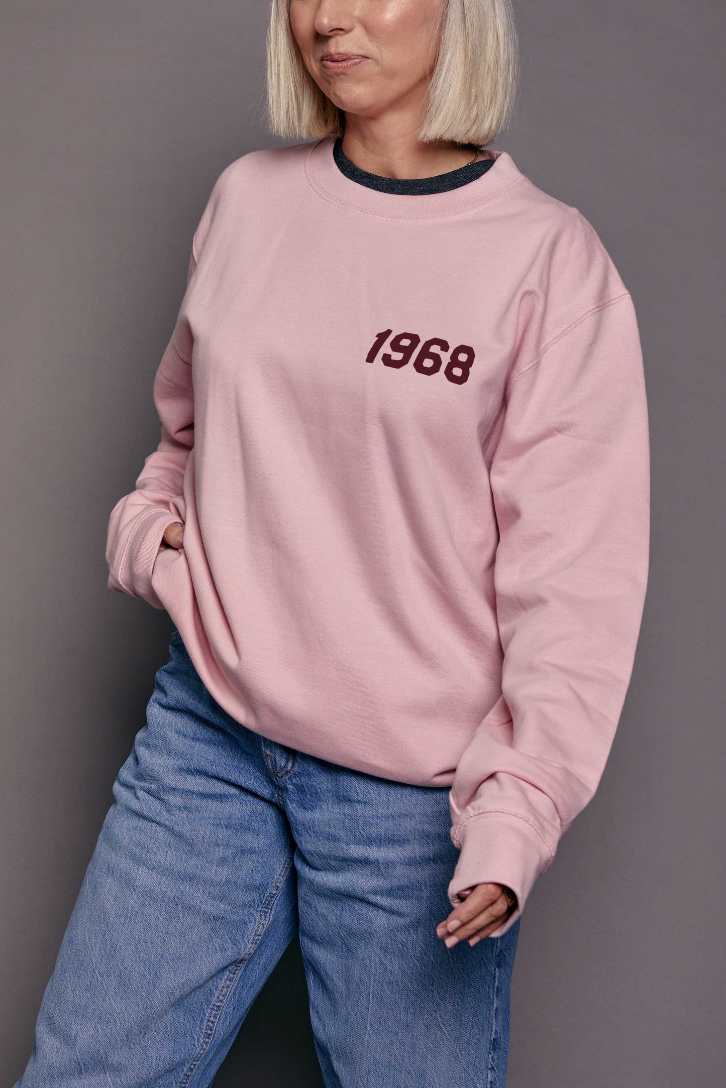 Person wearing a pink sweatshirt with '1968' printed on it in burgundy stood against a grey background