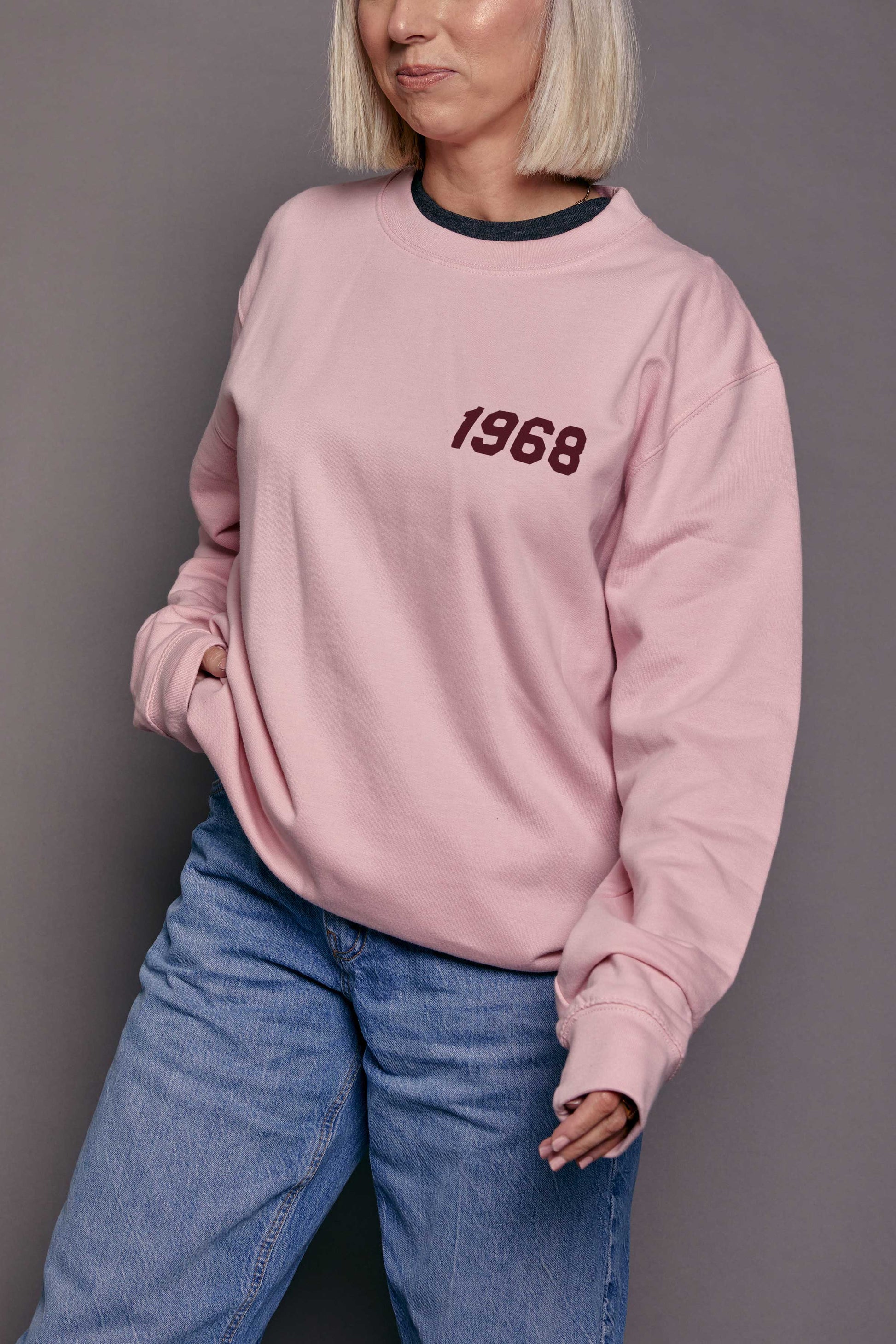 Person wearing a pink sweatshirt with '1968' printed on it in burgundy stood against a grey background