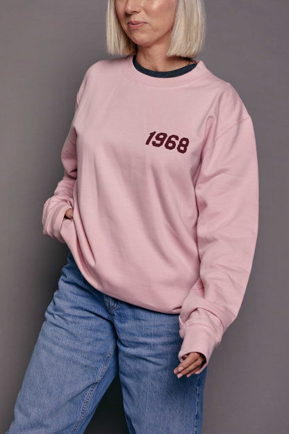 Person wearing a pink sweatshirt with '1968' printed on it in burgundy stood against a grey background