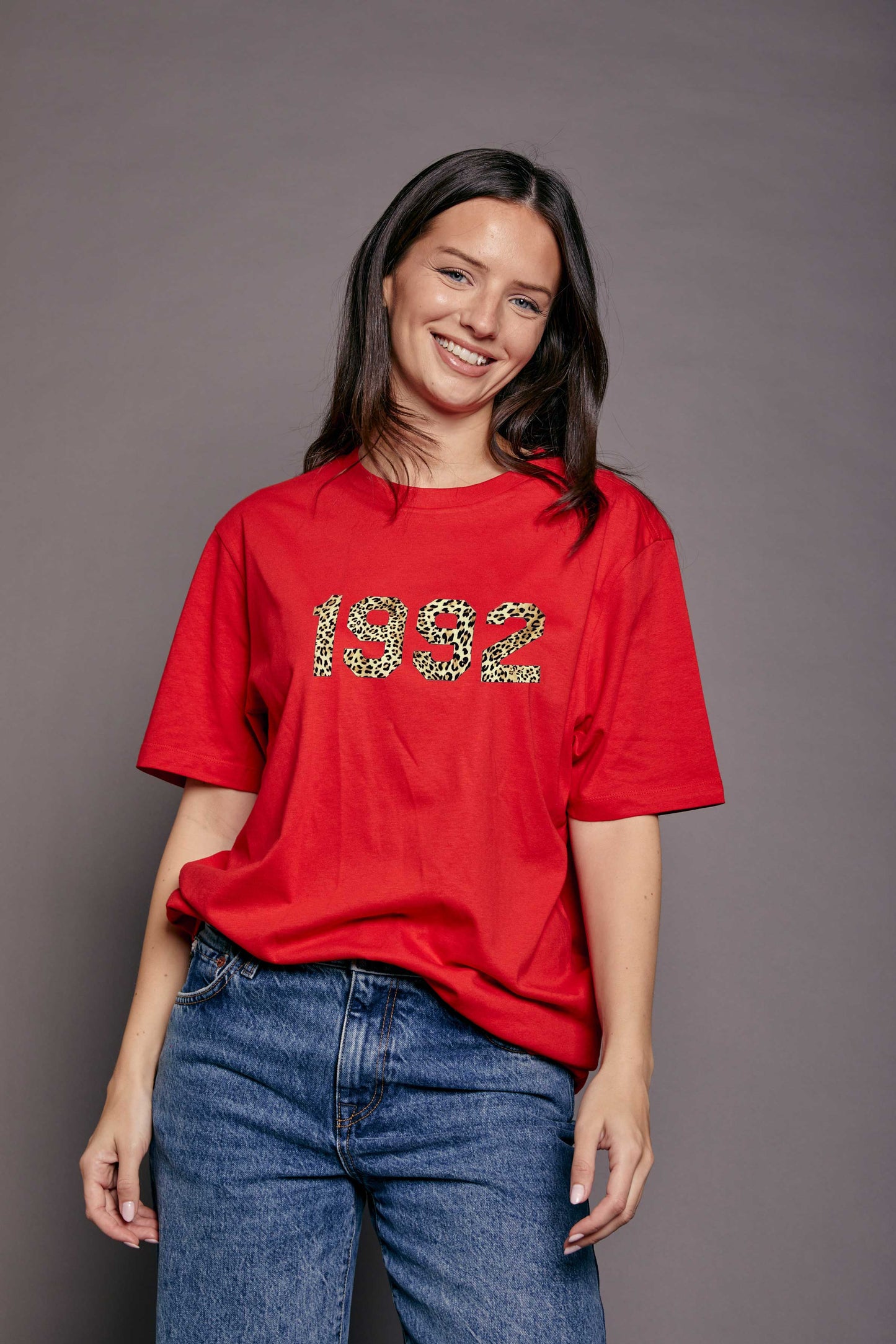 Woman wearing a red t-shirt with '1992' printed on it in leopard print stood against a grey background