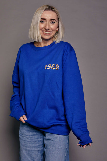 Person wearing a royal blue sweatshirt with '1968' printed in leopard print text stood in front of a grey background