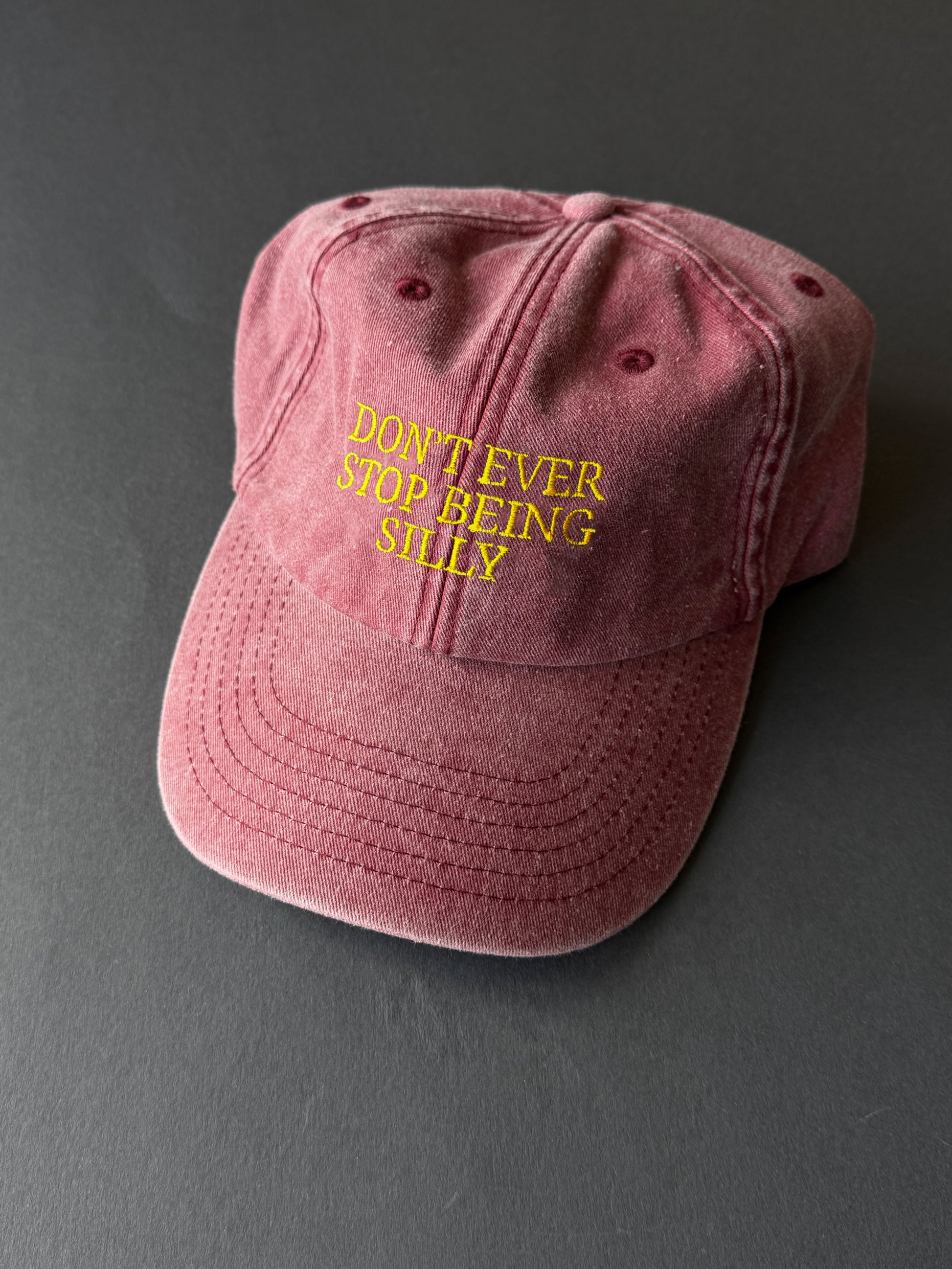 A washed red baseball cap with yellow embroidered text reading 'DON'T EVER STOP BEING SILLY' on the front, shown on a dark gray background.