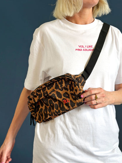 A person wearing a white t-shirt and blue plaid pants carries a leopard print crossbody bag with a black strap against a dark blue-grey background.