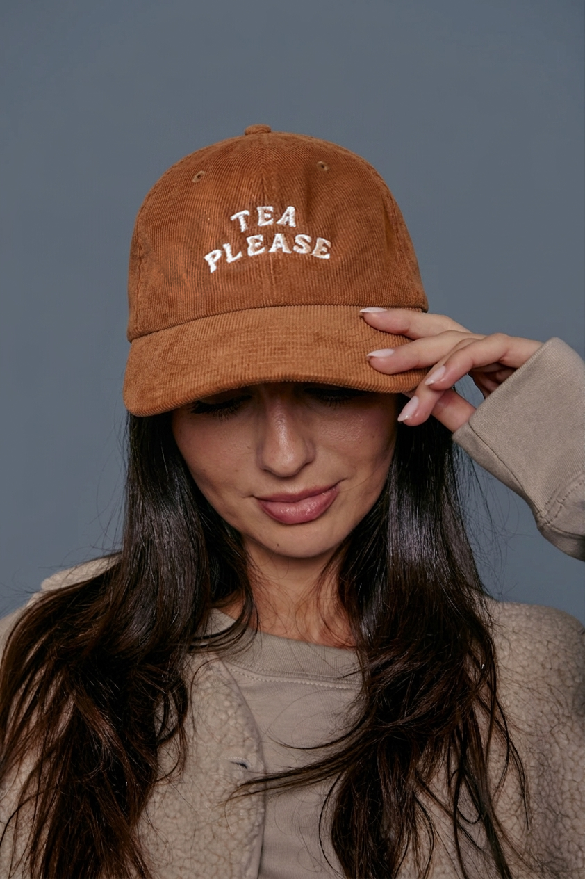 Person wearing a light brown cap with 'TEA PLEASE' text against a gray background