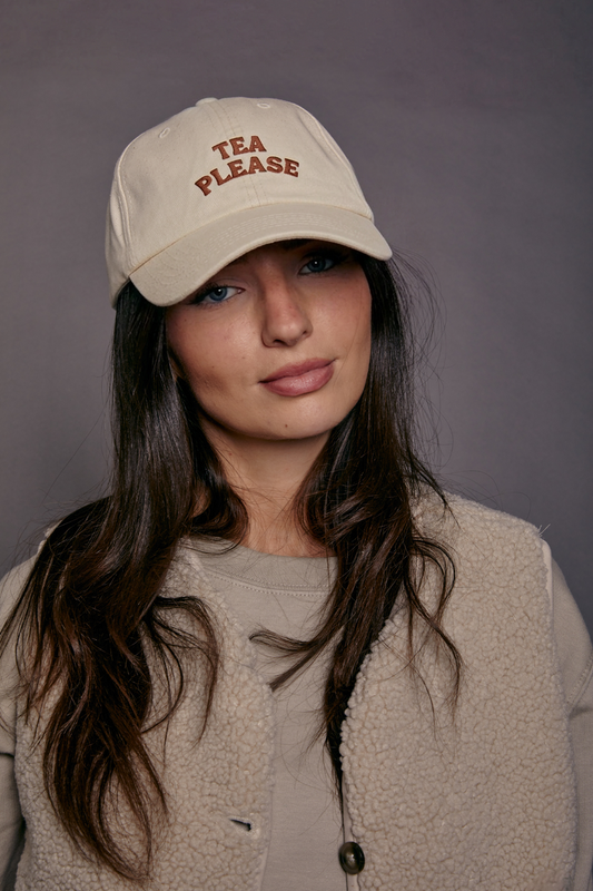 Woman wearing a beige cap with 'TEA PLEASE' text against a gray background