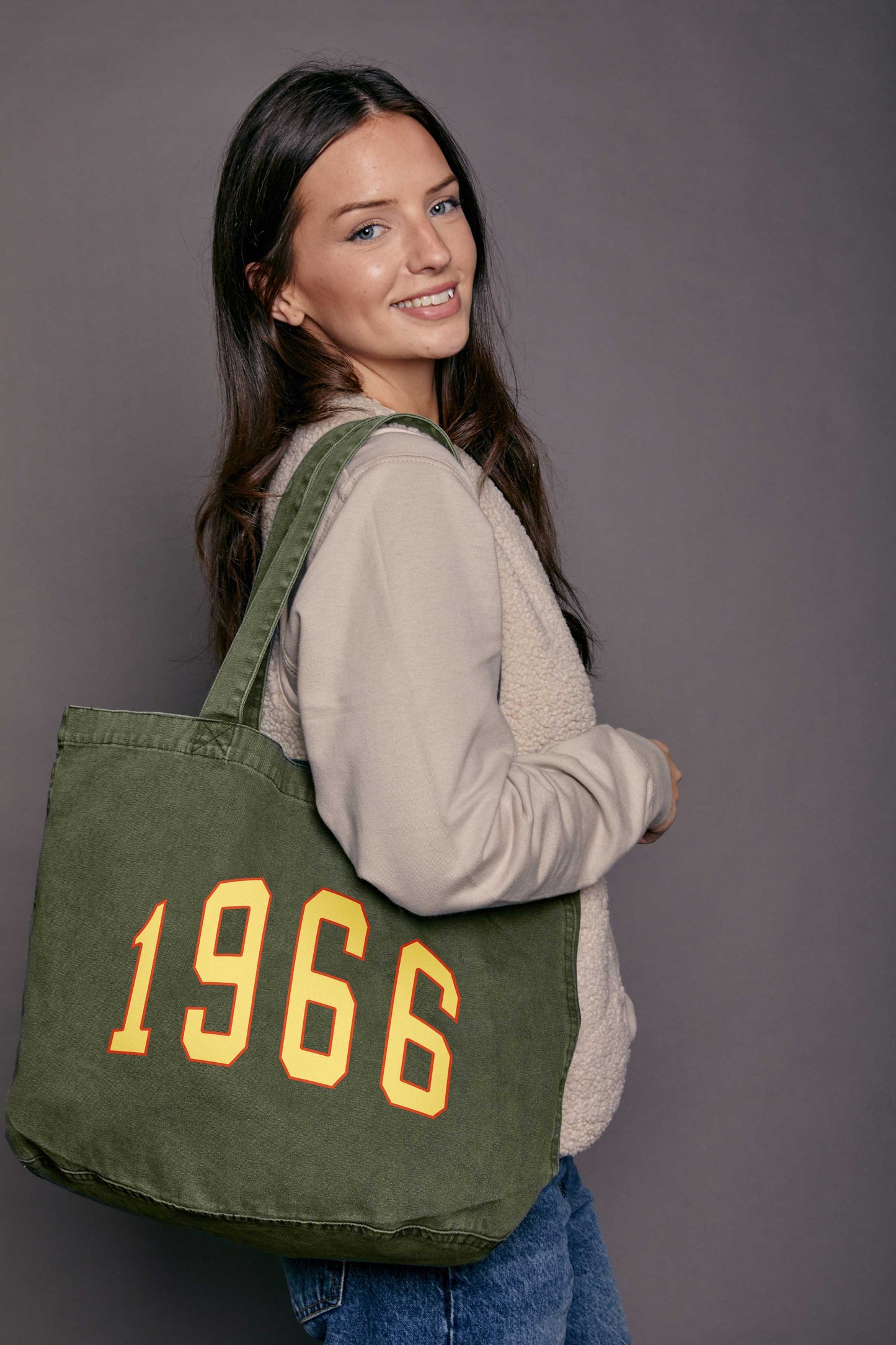 Woman carrying a khaki green tote bag with '1966' printed on it in yellow against a grey background