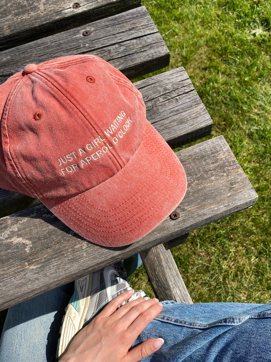 A vintage style, faded orange baseball cap with the embroidered slogan 'Just a girl waiting for Aperol O'clock' on a wooden bench.