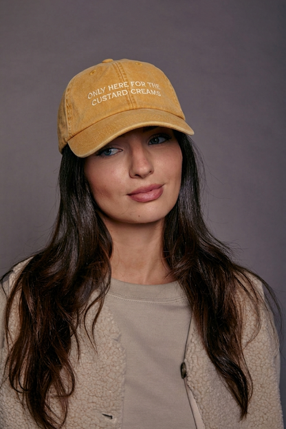 Woman wearing a yellow cap with text 'only here for the custard creams' against a grey background