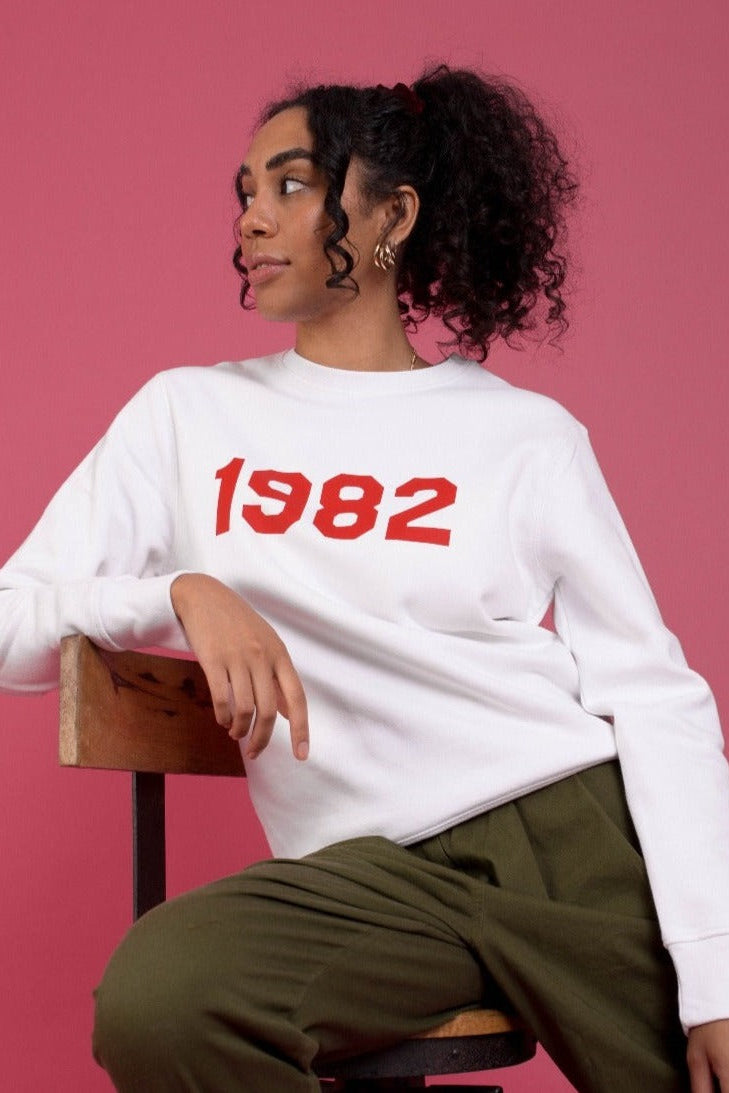 woman sat in front of pink background wearing a white, relaxed fit jumper printed with red text across the chest that reads "1982"