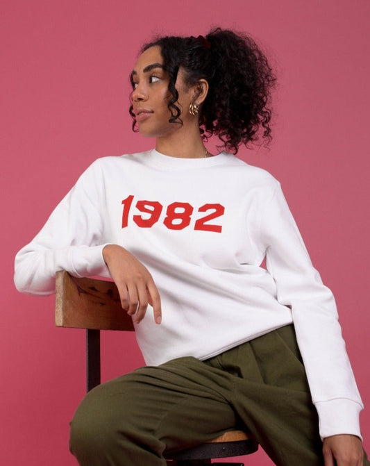 woman sat in front of pink background wearing a white, relaxed fit jumper printed with red text across the chest that reads "1982"