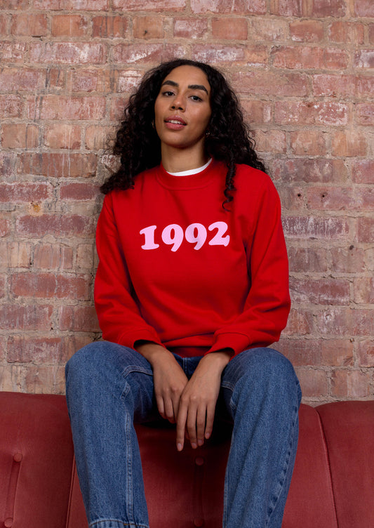 A person sitting on a couch wearing a red sweatshirt with the year '1992' printed in baby pink.